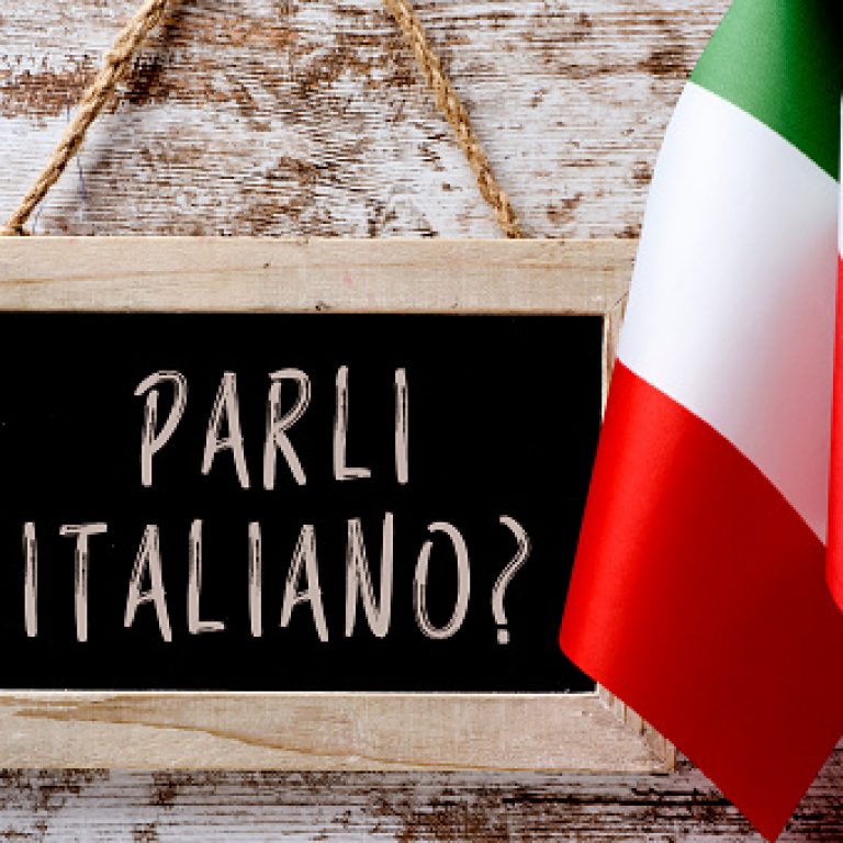 a wooden-framed chalkboard with the question parli italiano? do you speak Italian? written in Italian, and some flags of Italy against a rustic wooden background